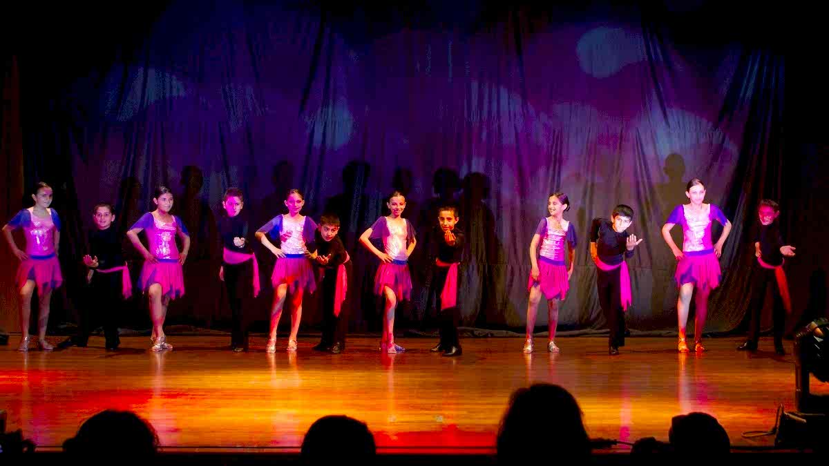Six couples of children, each couple paired as a girl and a boy, on a stage at an indoors dance recital.
